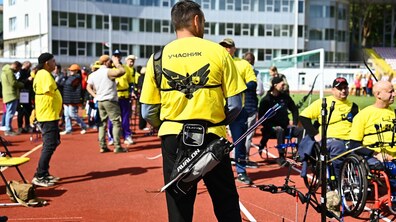 Спортивні змагання зі стрільби з лука, що проходять просто неба на стадіоні з біговими доріжками. На передньому плані спиною стоїть чоловік у жовтій футболці з написом «Учасник», на поясі якого закріплений сагайдак зі стрілами. На задньому плані та поруч видно інших учасників заходу, зокрема людей на кріслах колісних.