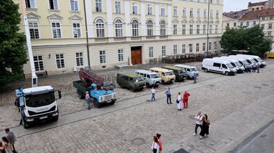 На передньому плані зліва стоїть велика сучасна біла вантажівка IVECO з піднятою довгою білою стрілою автовишки. Поруч із нею знаходиться старий блакитний вантажний автомобіль ГАЗ, в кузові якого закріплена складена дерев’яна драбина. Далі в цьому ж ряду вишикувано п'ять старих УАЗів різних відтінків: сірого, бежевого та зеленого кольорів. Далі стоять п'ять нових однакових білих мікроавтобусів FIAT Ducato.