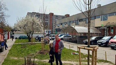 На фото пані Оля і її донька Тетянка стоять на зеленій ділянці біля нещодавно висаджених молодих дерев, які підпирають дерев’яні опори. На задньому плані видно житлові будинки