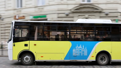 Автобус середнього класу рухається брукованою дорогою на тлі світлої кам’яниці. Його бічна частина пофарбована у яскраво-жовтий колір із великою блакитною вставкою та логотипом Львова по центру.
