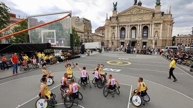 Змагання з баскетболу на кріслах колісних на площі перед Оперним театром у Львові: гравці двох команд у жовто-чорній та біло-червоній формах змагаються за м’яч, а навколо майданчика зібралися численні глядачі. На задньому плані видно Оперний театр.
