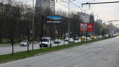 Панорама вулиці Стрийської на виїзді з міста. Дорога має кілька смуг у кожен бік, але рух тут не дуже щільний: видно окремі автомобілі та мікроавтобуси, які їдуть у напрямку до центру міста. Праворуч — велика порожня смуга дороги з потрісканим асфальтом і розміткою. Лівіше — розділювальна зелена зона з травою та невисокими деревами. Над дорогою тягнеться мережа тролейбусних дротів, закріплених на стовпах. На задньому плані — багатоповерхові житлові будинки сірого кольору