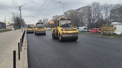 Частина вул. Стрийської під час робіт з асфальтування. Асфальт виглядає новим, рівним і ніби його щойно покладеним. По дорозі рухаються три великі дорожні катки — жовті машини для ущільнення асфальту. Вони їдуть один за одним у колоні, займаючи праву частину дороги. Ліворуч — тротуар, відділений від дороги низьким бордюром і рядом чорних металевих стовпчиків. Над дорогою видно дроти для тролейбусів, підвішені між стовпами.