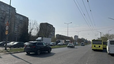 Вже відремонтована частина вул. Стрийської: дорога асфальтована, рівна, із промаркованими смугами для руху транспорту. На передньому плані видно автомобілі. Посередині вулиці є зелена розділювальна смуга з травою, яка відділяє зустрічні потоки транспорту. Уздовж дороги тягнуться високі стовпи з ліхтарями та тролейбусними дротами.