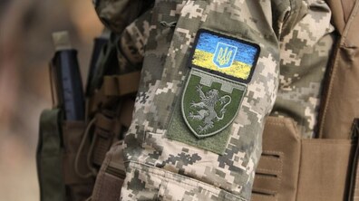 Плече військового у камуфляжній формі: на рукаві є шеврон із Прапором України та державним гербом – Тризубом; нижче розташований нарукавний знак військової частини – зелений щит із зображенням лева. Поруч також видно елементи тактичного спорядження військового.