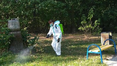 Жінка у спеціальному захисному костюмі білого кольору з розпилювачем проводить обробку зеленої ділянки в одному з парків міста. Поруч видно лавки та прогулянкові зони. Довкола ростуть зелені дерева та кущі