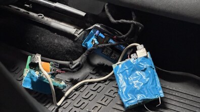 На фото видно розібране електронне обладнання (ймовірно, саморобний прослуховувальний пристрій), що лежить на рельєфному підлоговому килимку автомобіля. На відкритих частинах плат пристрою помітно мікросхеми, конденсатори, роз'єми та інші радіодеталі.