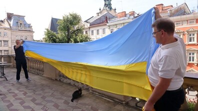 На відкритому балконі мер Львова Андрій Садовий і представник компаії «Ензим» розгортають великий синьо-жовтий прапор України. Під розгорнутим полотнищем підлозі балкона сидить смугастий кіт Левчик.