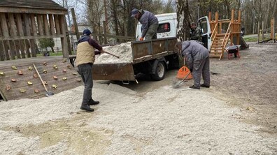 Троє комунальних працівників вивантажують пісок з машини. Один працює зверху на причепі вантажівки, інший інструментом забирає з причепа та розкидає на пісочницю дитячого майданчика, а третій з граблями вирівнює його рівномірно по території майданчика. Довкола чоловіків ігрові дерев’яні елементи дитячого майданчика: гірки та лазанки, а під ногами – пісок. Довкола ростуть дерева
