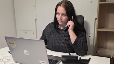 Дівчина сидить за столом і тримає в руці слухавку стаціонарного телефону. Перед нею ноутбук. Позаду - шафа