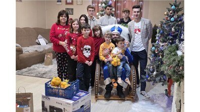 У центрі сидить Святий Миколай, а на колінах у нього двоє маленьких хлопчиків із подарунками в руках. Навколо стоять усміхнені діти та дорослі у різдвяних светрах. Праворуч видніється прикрашена ялинка, а попереду — коробки з подарунками та фруктами.