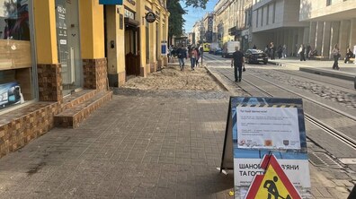 Ділянку вулиці Дорошенка, де проводять ремонт тротуару та облаштування доступних входів. Праворуч на передньому плані встановлено білий інформаційний стенд із попереджувальним дорожнім знаком «Дорожні роботи». Ліворуч видно розкопаний тротуар засипаний піском.