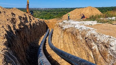 Масштабні роботи з прокладання нового колектора. Через відкриту піщану траншею великої глибини проходять дві великі чорні труби, які тягнуться вдалину. Біля краю розкопу працює екскаватор, поруч стоять двоє працівників у спецодязі. Навколо насипані високі пагорби ґрунту, а на задньому плані відкривається панорама зелених полів і околиць Львова.