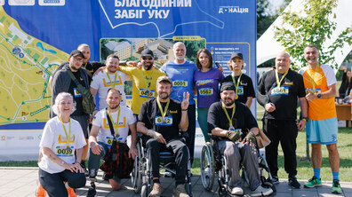 Учасники Першого благодійного забігу УКУ, які зібралися біля інформаційного стенда заходу. Серед учасників — ветерани, люди на кріслах колісних, військові та цивільні, які разом долучилися до благодійної ініціативи. Багато з них мають медалі та стартові номери, а дехто — протези чи засоби реабілітації.