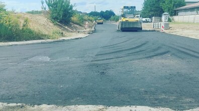 Ремонт вул. Конюшинної: дорожня техніка вкладає нижній шар асфальту на широкій ділянці дороги, а позаду працює дорожній коток. Обабіч вулиці видно насипи ґрунту, будівельні матеріали та сигнальні стовпчики.