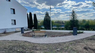 Облаштований простір із алеями, лавками та флагштоком. Поруч із доріжками — ділянки відкритого ґрунту для подальшого озеленення, а збоку видно білу будівлю та дерева