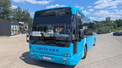 Світло-синій автобус №67, який стоїть на узбіччі дороги. На тлі видно дерева, кілька автомобілів і невелику будівлю.