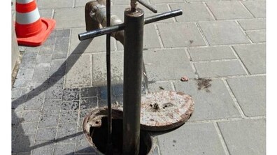 відкритий невеликий люк пожежного гідранта у тротуарній плитці. З отвору люка вертикально вгору піднімається високе металеве обладнання, для ремонту. Поруч стоїть дорожній конус, що сигналізує про проведення робіт.