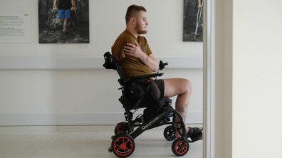 У холі центру UNBROKEN ветеран із ампутованими рукою та ногою сидить у спеціальному кріслі колісному: він повернутий у профіль і тримає ліву руку на грудях так, наче закриває місце ампутації правої руки. Він дивиться вперед із дуже зосередженим та замисленим виразом обличчя. На стіні позаду видно фотографії людей із допоміжними засобами для пересування, що підкреслює тему реабілітації.