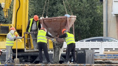 Будівельний майданчик шляхопроводу. Видно процес бетонування: робітники в яскравих жилетах і помаранчевих касках спрямовують підвішену на крані ємність із бетоном в опалубку конструкції.