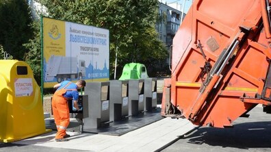 Контейнерний майданчик для роздільного збору відходів. Ліворуч стоїть жовтий контейнер для пластику, поруч працівник комунальної служби у спецодязі проводить технічне обслуговування обладнання. У центрі розміщені контейнери для різних видів відходів, а праворуч видно частину сміттєвоза. На задньому плані — житловий будинок і зелень.
