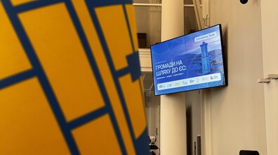 На великому екрані телевізора, який висить на стіні на синьому тлі білими літерами написано – Громади на шляху до ЄС. Позаду велика біла колона. На передньому плані стяг з синіх та жовтих кольорів з візерунками