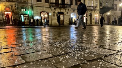 Вечірня вулиця старого міста, вимощена мокрою бруківкою, на якій розсипані дрібні крижинки мокрого снігу. На задньому плані видно силуети перехожих та історичні будівлі з підсвіченими вікнами.