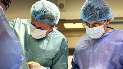 Двоє медичних працівників під час операції: одягнені в хірургічні халати, маски, рукавички та одноразові шапочки, зосереджено дивляться вниз (на операційне поле). Один із них тримає хірургічний інструмент, інший – допомагає