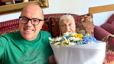 Ювілярка Іванна Скарачинська-Димнич та працівник управління соціального захисту Іван Фасольник усміхаються, позуючи для спільного селфі. Пані Ніна тримає в руках великий букет різнокольорових квітів.