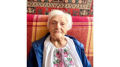 Ювілярка Олена Рибалко з лагідним і спокійним поглядом сидить на дивані, вкритому яскравим картатим пледом. Вона одягнена у традиційну вишиванку з рожевими квітковими орнаментами та синю кофту. За її спиною видно декоративний килим з візерунками.