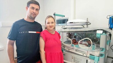 Чоловік та жінка стоять поруч із медичним інкубатором для новонароджених. Вони дивляться в камеру й усміхаються, а біля них видно дитину, яка перебуває під наглядом у лікарняній палаті.