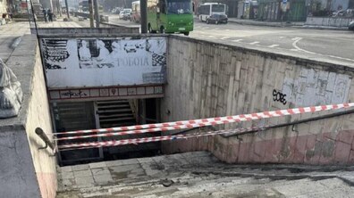 Вхід до підземного переходу. Сходи та пандус перекриті червоно-білою стрічкою, натягнутою поперек проходу. Стіни переходу облицьовані світлою плиткою, місцями видно графіті. На задньому плані — міська вулиця: їде зелений автобус, кілька автомобілів, вздовж дороги стоять стовпи та будівлі.