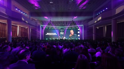 Дискусія під час конференції IT Arena 2026, у якій бере участь міський голова Андрій Садовий. У залі зібралося багато людей, які сидять спиною до камери та дивляться на сцену. Сцена оснащена великими екранами та яскравим фіолетово-зеленим освітленням із прожекторами.