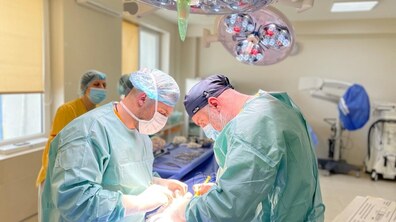 Двоє лікарів у захисному одязі, масках і шапочках оперують в операційній. Над ними яскраво світять дві великі операційні лампи. Позаду видно медичну сестру та різне медичне обладнання й інструменти.