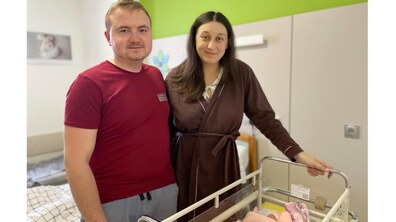 Усміхнені Ганна та Василь стоять у лікарняній палаті після народження дитини. Чоловік у червоній футболці стоїть зліва, жінка у коричневому халаті – справа. Поруч із ними стоїть прозоре дитяче ліжечко на колесах, у якому лежить немовля, загорнуте в рожеву ковдру.