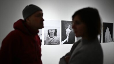 Інтер’єр художньої галереї з білою стіною. На стіні висять кілька чорно-білих фотографій, розташованих у ряд. На одній з них видно руку, що тримає маленьке дзеркало або рамку з відображенням нижньої частини жіночого обличчя на тлі води. На передньому плані — дві людини, зняті навмисно нечітко, у напівтемряві.