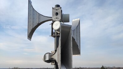 Велика металева сирена оповіщення, розташована просто неба на тлі хмарного неба та житлової місцевості. Вона має кілька рупорів, спрямованих у різні боки для поширення звукового сигналу на значну відстань.