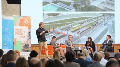 Виступ Антона Коломєйцева на Urban Mobility Forum 2026 - головний архітектор Львова стоїть ліворуч на сцені в чорній сорочці та бежевих штанах і активно жестикулює під час промови. Праворуч від нього в кріслах сидять ще чотири учасники панельної дискусії. За їхніми спинами розташований великий екран із презентацією архітектурних та інфраструктурних проєктів міського простору.
