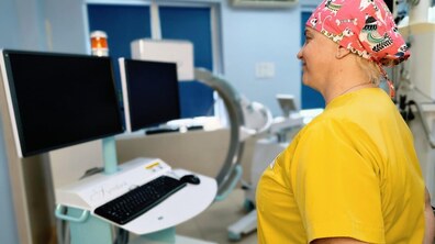 Медична працівниця в жовтій формі та медичній шапочці з кольоровими собаками стоїть в кабінеті поруч з мобільним цифровим рентгенхірургічним апаратом. Перед нею сучасні монітори