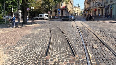 Перехрестя вулиць Князя Романа – І. Білозіра – Івана Франка з трамвайними коліями, вимощене старою бруківкою. Одна трамвайна колія обірвана. Навколо кілька автомобілів, мотоцикліст, велосипедисти і пішоходи, а обабіч вулиці стоять багатоповерхові історичні будинки та великі дерева.
