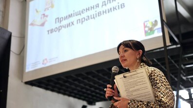 Очільниця управління комунальної власності ЛМР Наталія Рясна виступає перед митцями. Позаду неї видніється проєктор з презентацією