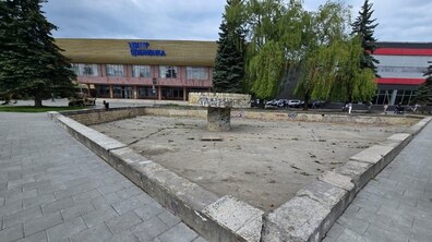Площа перед будівлею «Центру Довженка». На передньому плані видно стару конструкцію із потрісканим бетонним дном та невеликою спорудою посередині, розмальованою графіті. Навколо сіра плитка пішохідної зони. Позаду розташована велика будівля культурного центру з написом «Центр Довженка» синіми літерами на фасаді. Праворуч і ліворуч ростуть високі дерева, зокрема велика плакуча верба.