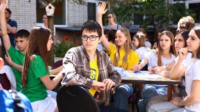 Діти сидять за столом на подвір’ї школи. Дехто підняв руку.