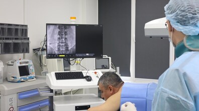 У медичному кабінеті лікар проводить процедуру пацієнту. На лікарняному ліжку лежить чоловік. Поруч стоїть медик у спецодязі. На екрані попереду видно хребет пацієнта. В кабінеті медичні прилади для обстежень та процедур
