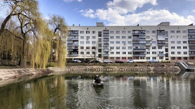Комунальні працівники на невеликому човні посеред невеликого озера. Збоку поруч з водоймою похилились верби. Позаду озера шестиповерховий житловий будинок