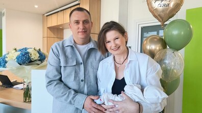 Пацієнтка Марія з чоловіком Олегом та сином Яремою стоять у холі дитячої лікарні святого Миколая. Марія тримає на руках сина, а поруч видно повітряні кульки та букет блакитно-білих квітів.
