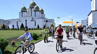 Учасники велопрощі – дорослі та діти їдуть на велосипедах широкою доріжкою. Позаду видно великий сучасний храм – церкву Різдва Пресвятої Богородиці на Сихові, та український прапор.