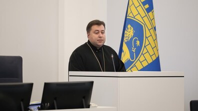 Отець Тарас Михальчук у чорному священичому одязі стоїть за трибуною у великій сесійній залі Львівської міської ради. Позаду видно великий прапор міста.