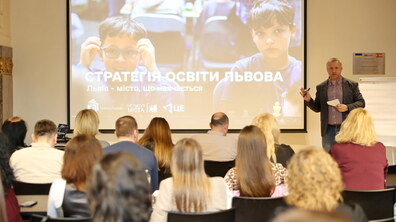 У світлій аудиторії проходить презентація «Стратегії освіти Львова». На великому екрані зображені двоє школярів, а перед аудиторією виступає керівник стратегічних програм Інституту міста Андрій Пундор, пояснюючи ключові тези слухачам, що сидять спиною до камери.