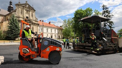 Дорожні робітники кладуть новий асфальт на вул. Винниченка біля церкви. На передньому плані — помаранчевий дорожній каток із водієм у яскравому жилеті, праворуч — велика асфальтоукладальна машина та кілька працівників. На тлі видно історичні будівлі, дерева й темні хмари з просвітами блакитного неба.